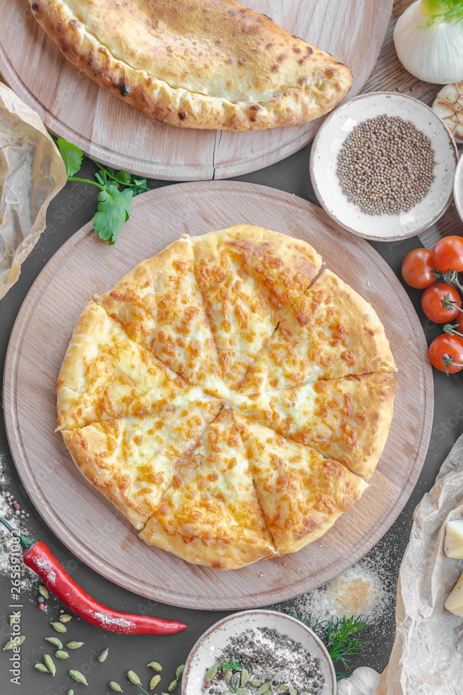 Georgian khachapuri pie with cheese on wooden plate, cheese, red pepper, tomatoes, mushrooms, herbs, onion and bowls with spices nearby on the table. Horizontal image. Top view, flat lay. Georgian khachapuri pie with cheese on wooden plate, cheese, red pepper, tomatoes, mushrooms, herbs, onion and bowls with spices nearby on the table. Horizontal image. Top view, flat lay.