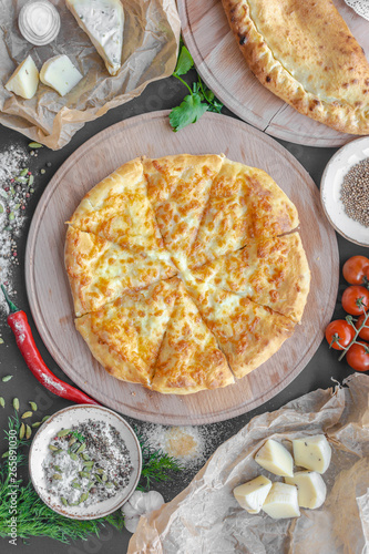 Georgian khachapuri pie with cheese on wooden plate, cheese, red pepper, tomatoes, mushrooms, herbs, onion and bowls with spices nearby on the table. Horizontal image. Top view, flat lay.