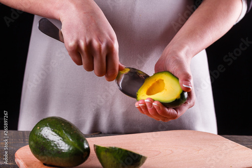 A young woman in an gray apron cuts an avocado on a wooden cutting board