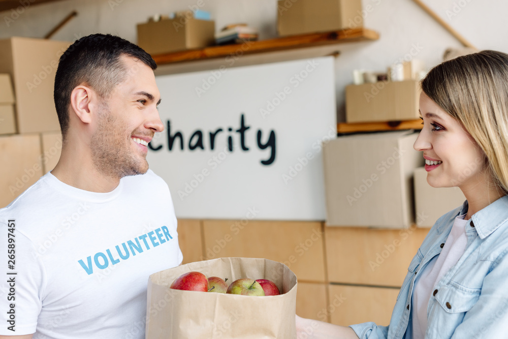 handsome, smiling volunteer giving paper bag with apples to attractive woman handsome, smiling volunteer giving paper bag with apples to attractive woman