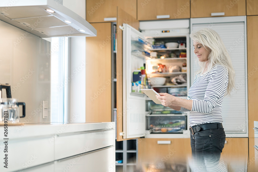 © Westend61 - Content mature woman standing in kitchen at smart home checking fridge with digital tablet