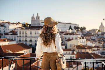 Blonde woman standing on the balcony and looking at coast view of the southern european city with sea during the sunset, wearing hat, cork bag, safari shorts and white shirt