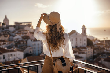 Blonde woman standing on the balcony and looking at coast view of the southern european city with sea during the sunset, wearing hat, cork bag, safari shorts and white shirt
