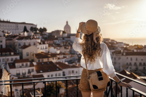 Blonde woman standing on the balcony and looking at coast view of the southern european city with sea during the sunset, wearing hat, cork bag, safari shorts and white shirt photo