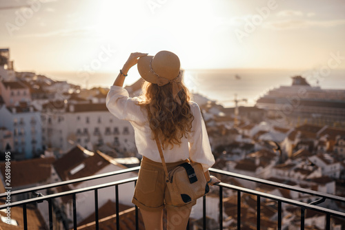 Blonde woman standing on the balcony and looking at coast view of the southern european city with sea during the sunset, wearing hat, cork bag, safari shorts and white shirt