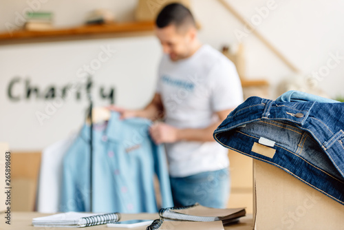 selective focus of young volunteer standing near rack with shirts, and cardboard box with jeans