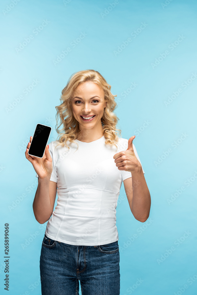 Obraz premium cheerful blonde woman showing thumb up while holding smartphone with blank screen on blue