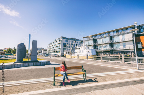 Reykjavik city lifestyle people watching street scene, Iceland. Woman using smart phone relaxing on bench at reykjavík harbour near modern condo building downtown. Summer Europe lifestyle.