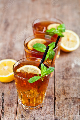 Cold iced tea with lemon, mint leaves and ice cubes in three glasses on rustic wooden table.
