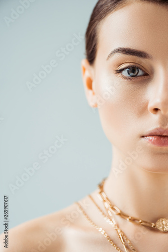 cropped view of young woman with shiny makeup in golden necklaces looking at camera isolated on grey
