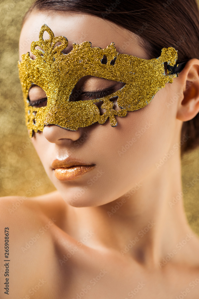 young naked woman in golden mask with closed eyes, shiny makeup looking away on textured golden background