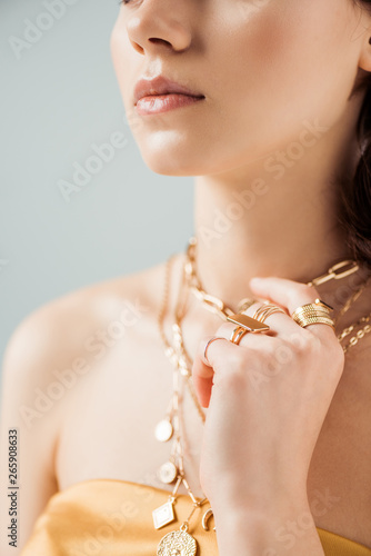 partial view of young woman with shiny lips in golden necklaces and rings isolated on grey