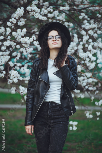 Outdoor spring portrait of fashion sensual young girl posing in amazing blooming garden. Wearing a hat and leather jacket  feminine look  elegant glamorous style. On the background of cherry blossoms