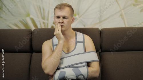 Painful, bored man with a broken arm wearing arm brace sitting on a sofa photo