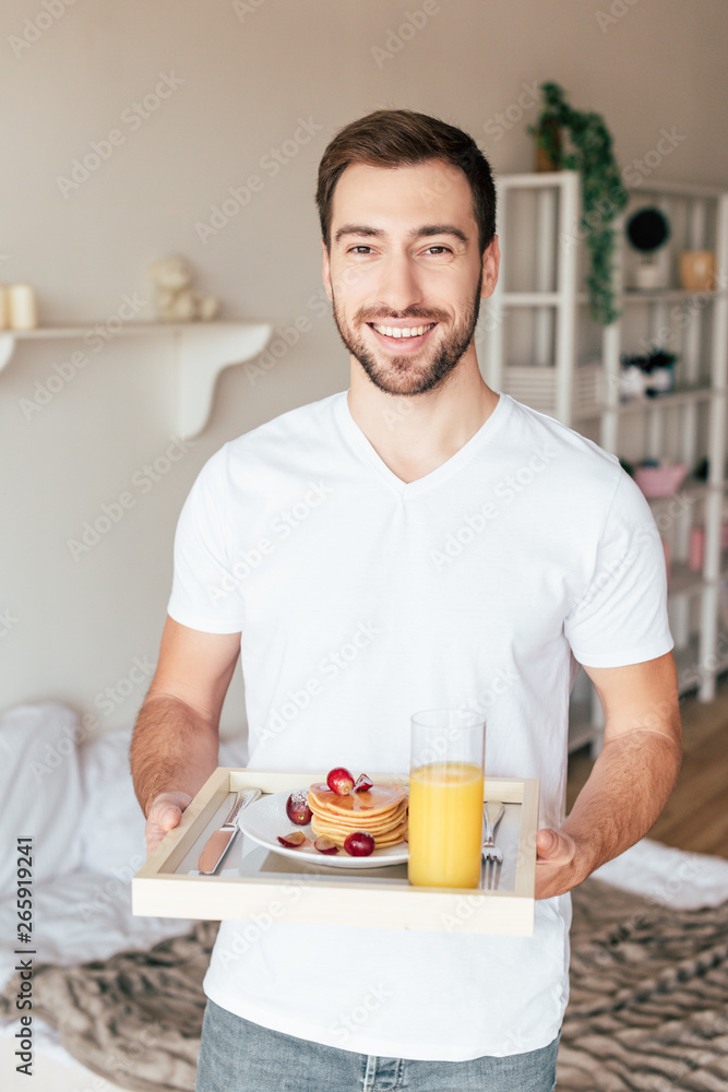smiling man holding tray with pancakes and glass of orange juice in bedroom smiling man holding tray with pancakes and glass of orange juice in bedroom