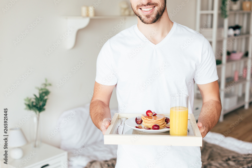 cropped view of smiling man holding tray with pancakes and glass of orange juice in bedroom cropped view of smiling man holding tray with pancakes and glass of orange juice in bedroom