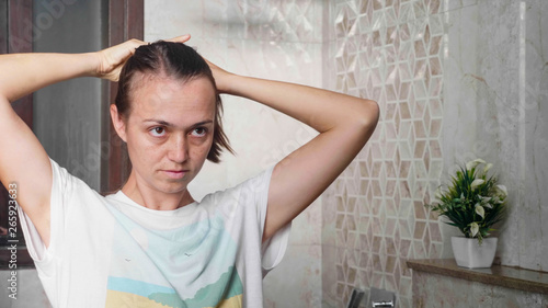 Young brown-haired woman plaits long silky hair at bathroom in front of mirror in ponytail. Portrait shot in mirror reflection.
