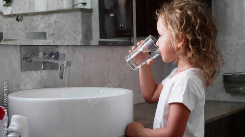 Slika na platnu Cute little curly child girl rinses her mouth with water, looking at mirror and spits water into the sink, at domestic bathroom, side view