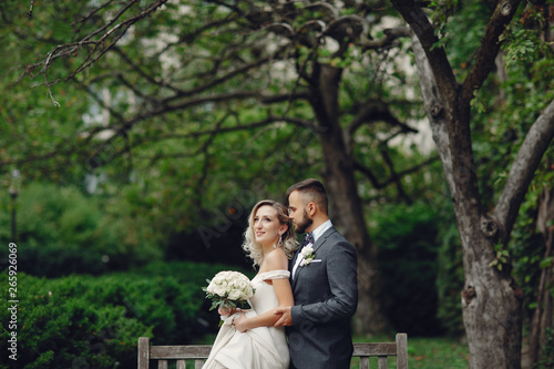 Beautiful bride in a long white dress. Handsome groom in a black suit. Couple in a summer park sitting on a bench