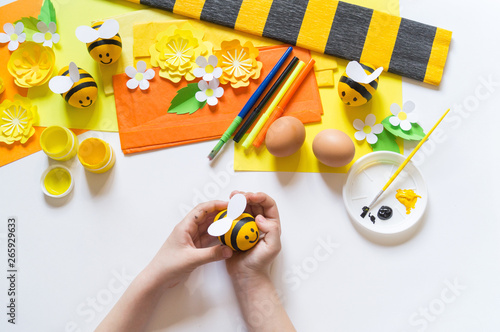 Child hands girl paints an Easter egg. Egg a yellow bee. White background. Material for creativity,