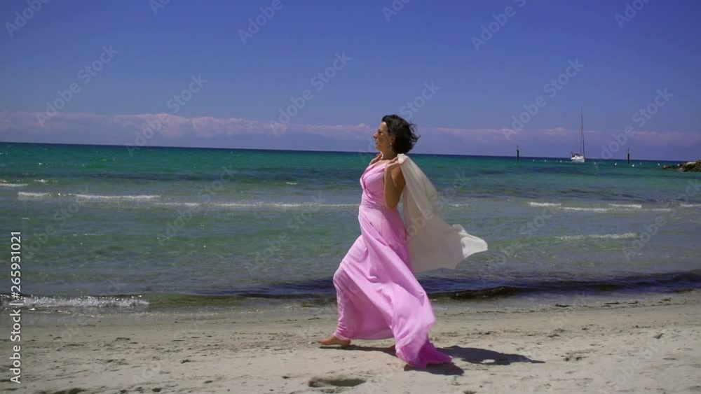 A dark-haired tanned woman in dark glasses and a long pink dress runs and circles along the sea on a sandy beach barefoot. Her dress and shawl flutter in the wind. A dark-haired tanned woman in dark glasses and a long pink dress runs and circles along the sea on a sandy beach barefoot. Her dress and shawl flutter in the wind.
