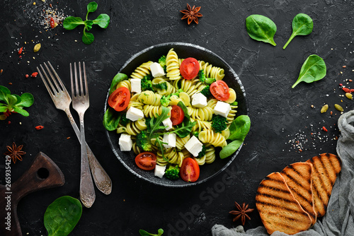 Pasta with spinach, tomatoes and feta cheese. In a black plate on a wooden background Top view. Free space for your text. Flat lay