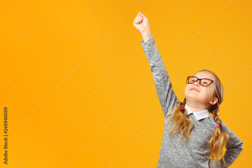 Closeup of little child girl on the background of yellow wall. Schoolgirl with glasses raised her hand to the top. The concept of success and education. Closeup of little child girl on the background of yellow wall. Schoolgirl with glasses raised her hand to the top. The concept of success and education.