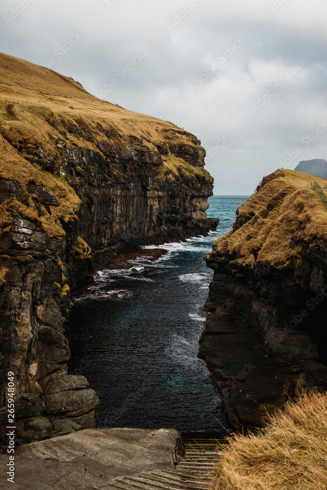 Direct view into the gorge / natural harbour of Gjógv with dramatic cloudy sky, lush orange grass and turquoise water (Faroe Islands, Denmark, Europe
