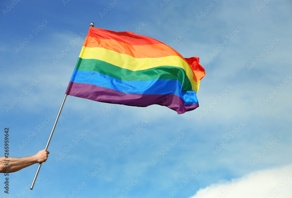 Gay man holding rainbow LGBT flag against blue sky Gay man holding rainbow LGBT flag against blue sky