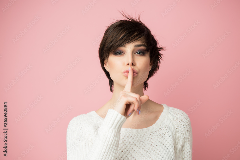 Beautiful young woman with long shot dark hair wears casual white sweater shows hush sign with serious expression. Pretty lady asks not to tell her secret anybody. Beautiful young woman with long shot dark hair wears casual white sweater shows hush sign with serious expression. Pretty lady asks not to tell her secret anybody.