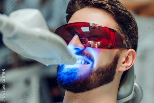 Man having teeth whitening by dental UV whitening device,dental assistant taking care of patient,eyes protected with glasses. Whitening treatment with light, laser, fluoride. photo