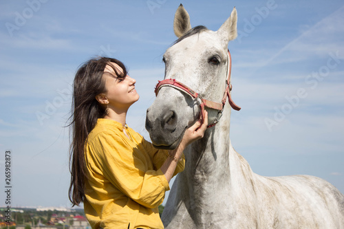 Beautiful young girl smiling and laughing, pretty european woman in yellow bright shirt hugging adorable white horse in the sunlight shine, with blue sky. Horse farm, horse care, horse riding activity