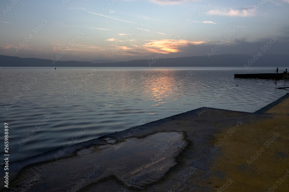 Sunset over Lake Ohrid from a pier with a puddle that reflects a trace of airplane.