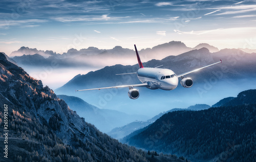 Fototapeta Naklejka Na Ścianę i Meble -  Airplane is flying over mountains in fog at sunset in summer. Landscape with passenger airplane, hills in low clouds, blue sky. White aircraft. Business travel. Commercial plane. Aerial view