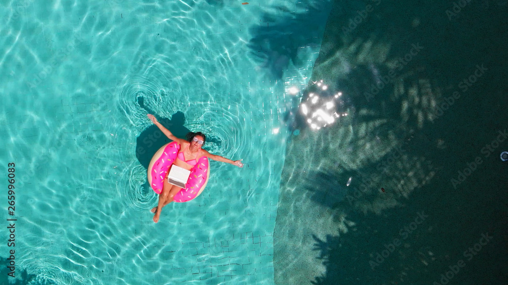 Aerial view of a young brunette woman swimming on an inflatable big donut with a laptop in a transparent turquoise pool.