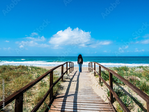 a wooden path leading to the beach, in the background a woman walking towards the sea, in the wild nature. Italian landscape