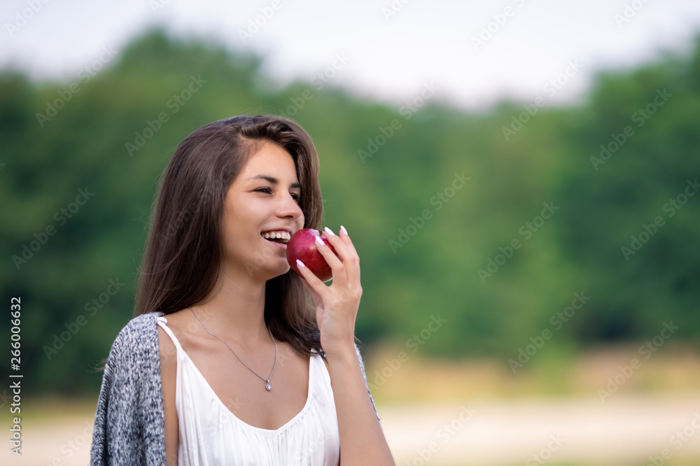 Young beautiful girl in white dress eating ripe organic apple. She is on the shore of the lake and enjoy a picnic and outdoor recreation.