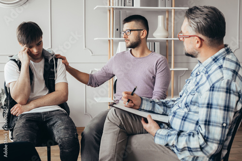 Young man in glasses comforting his depressed friend during meeting with counselor photo