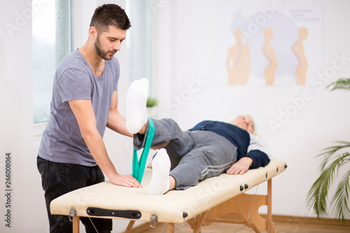Grey elderly man lies on a physiotherapy table, and young doctor helps him during exercises