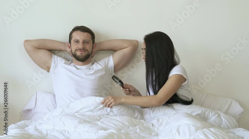 Couple lying in the bed. Girlfriend joking and checking potency using magnifying glass. Boyfriend smiling and looking at the camera.