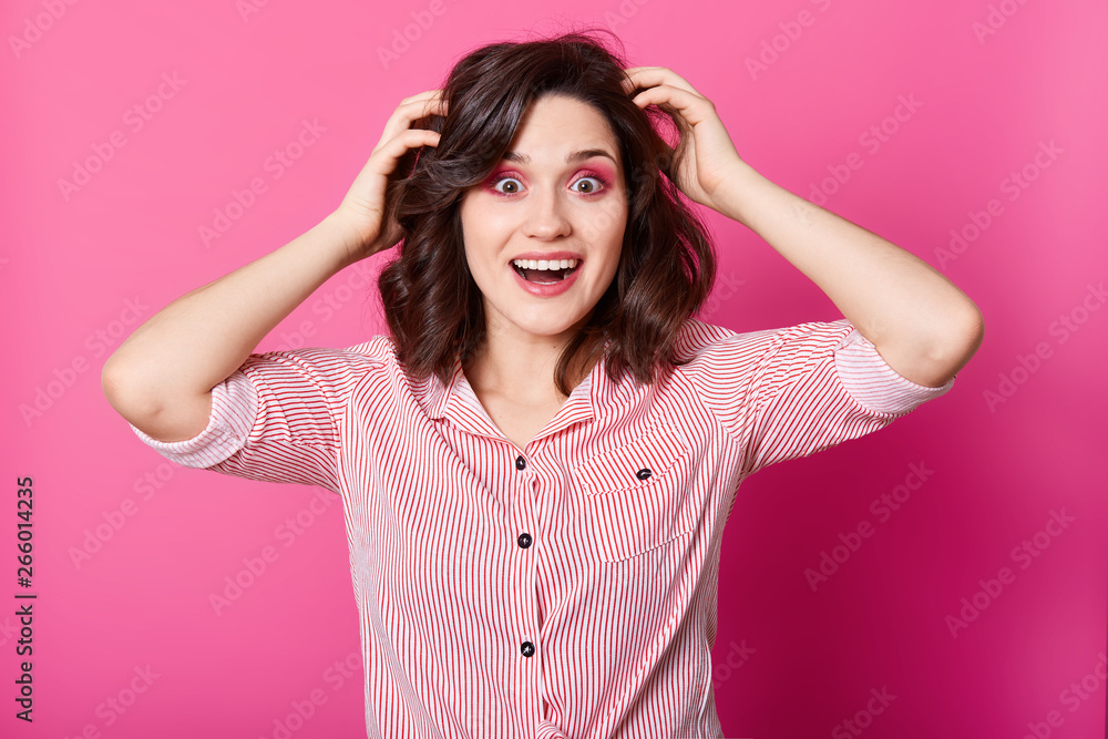 Close up portrait of beautiful dark haired woman holding hands on head with surprise, keeps eyes and mouth widely open, wearing stripped shirt, lady with happy facial expression. Human motions concept Close up portrait of beautiful dark haired woman holding hands on head with surprise, keeps eyes and mouth widely open, wearing stripped shirt, lady with happy facial expression. Human motions concept