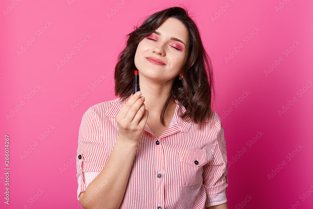 Half length photo of woman stands with closed eyes and holding red lipstick. Beauty portrait of model with dark wavy hair, dressed stylish blouse, poses in studio with rose background. Beauty concept. Half length photo of woman stands with closed eyes and holding red lipstick. Beauty portrait of model with dark wavy hair, dressed stylish blouse, poses in studio with rose background. Beauty concept.