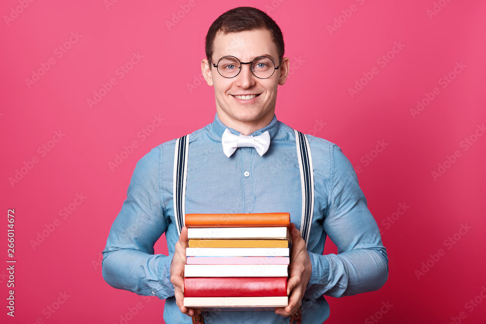 Smiling positive man holds bunch of colorful books in both hands, posing isolated over pink background in studio. Athletic strong man wears blue shirt, stylish spectacles, bow tie and suspenders. Smiling positive man holds bunch of colorful books in both hands, posing isolated over pink background in studio. Athletic strong man wears blue shirt, stylish spectacles, bow tie and suspenders.
