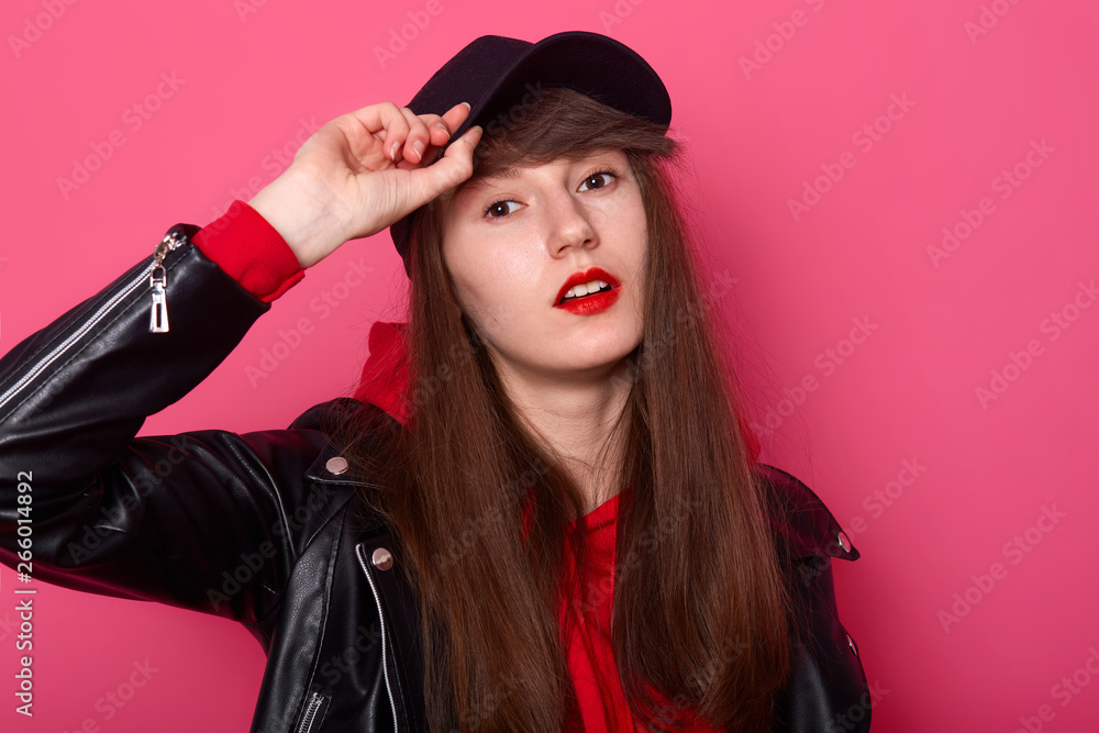 Indoor shot of happy Caucasian young teenager girl in red hoodie, leather jacket, stands with cheeky facial expression, keeps hand on black visor of cap against rose studio wall. People concept Indoor shot of happy Caucasian young teenager girl in red hoodie, leather jacket, stands with cheeky facial expression, keeps hand on black visor of cap against rose studio wall. People concept