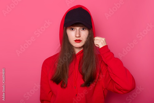 Image of brunette girl looking directly at camera with calm facial expression, wearing casual red hoody and black cap, touches her hood, posing isolated on rose background. Modeling concept.