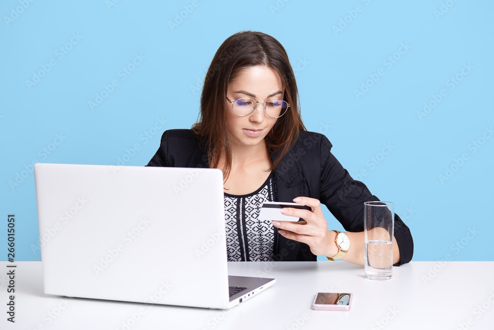 Attentive attractive young businessman sits at white desk, working with laptop, typing credit card information, wearing classic blouse, black jacket and fashionable spectacles. Office work concept. Attentive attractive young businessman sits at white desk, working with laptop, typing credit card information, wearing classic blouse, black jacket and fashionable spectacles. Office work concept.