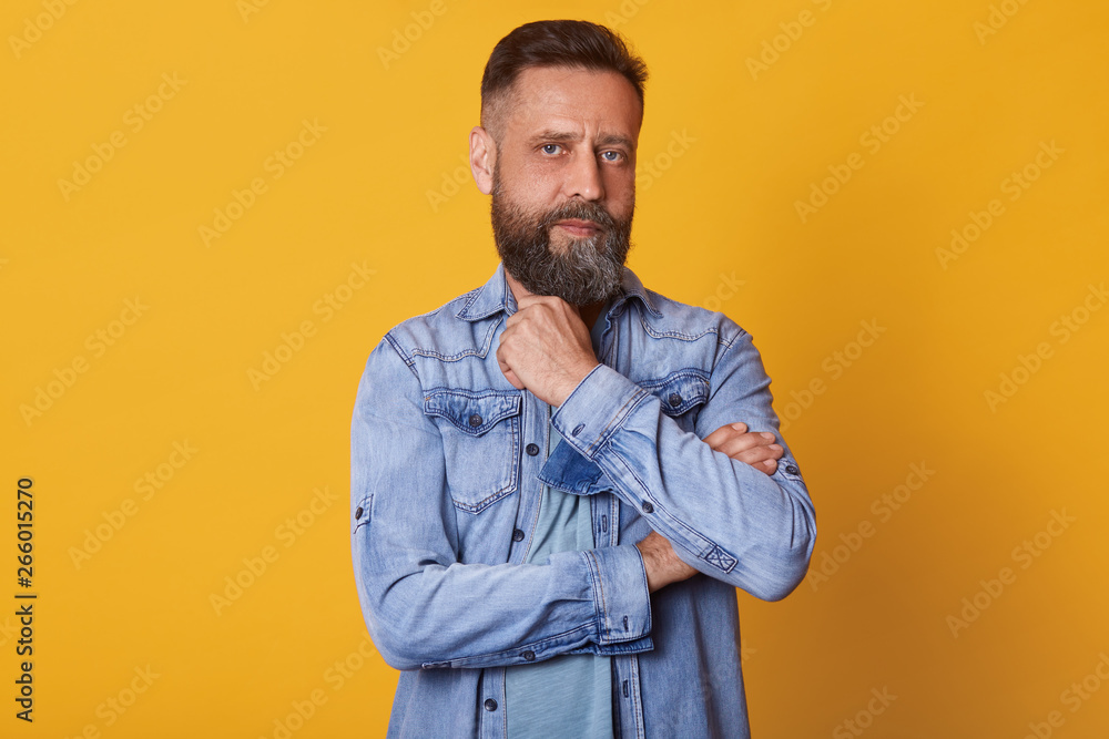 Strong thoughtful blue eyed man looking directly at camera, posing isolated over bright yellow background in studio, touching his beard with fingers, having powerful look, wearing in casual manner.