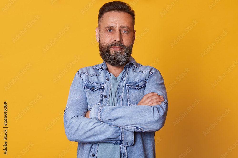 Fototapeta premium Close up portrait of bearded middle aged confident man with folded arms, standing straight, having serious facial expression, wearing casual jeans jacket, looks tired and determined. Emotions concept.