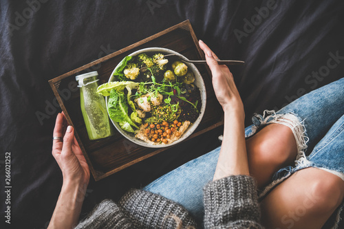 Healthy dinner, lunch in bed. Vegan superbowl or Buddha bowl with hummus, vegetable, fresh salad, beans, couscous and avocado, green smoothie on tray and woman in jeans eating with fork, top view