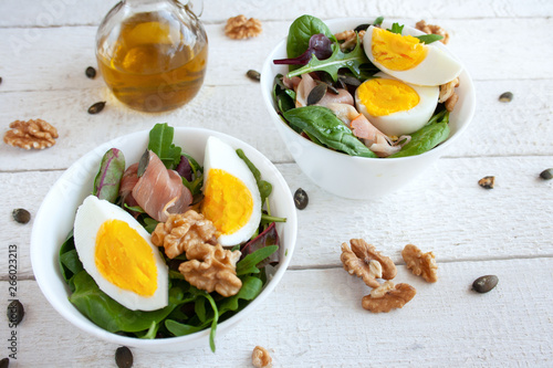 Salad with egg, walnuts and parma ham in white bowls, olive oil in the background